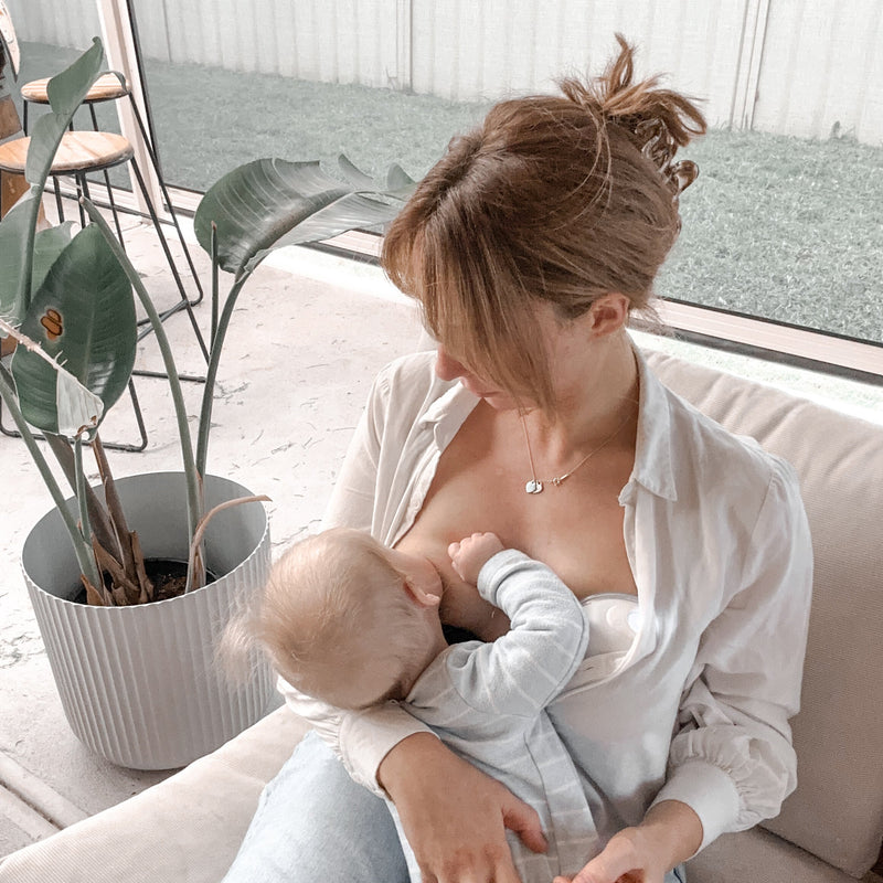 Woman breastfeeding a baby on a couch with a plant and window in the background